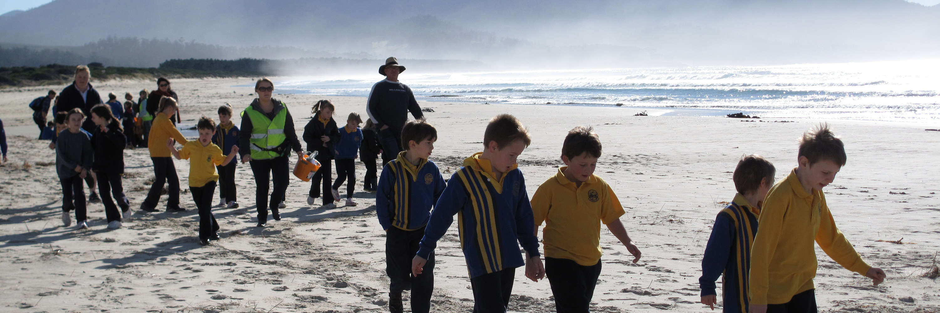 A line of young students and teachers walk along a white sandy beach with sunlit haze and mountains behind. Photo: Andrew Hughes.