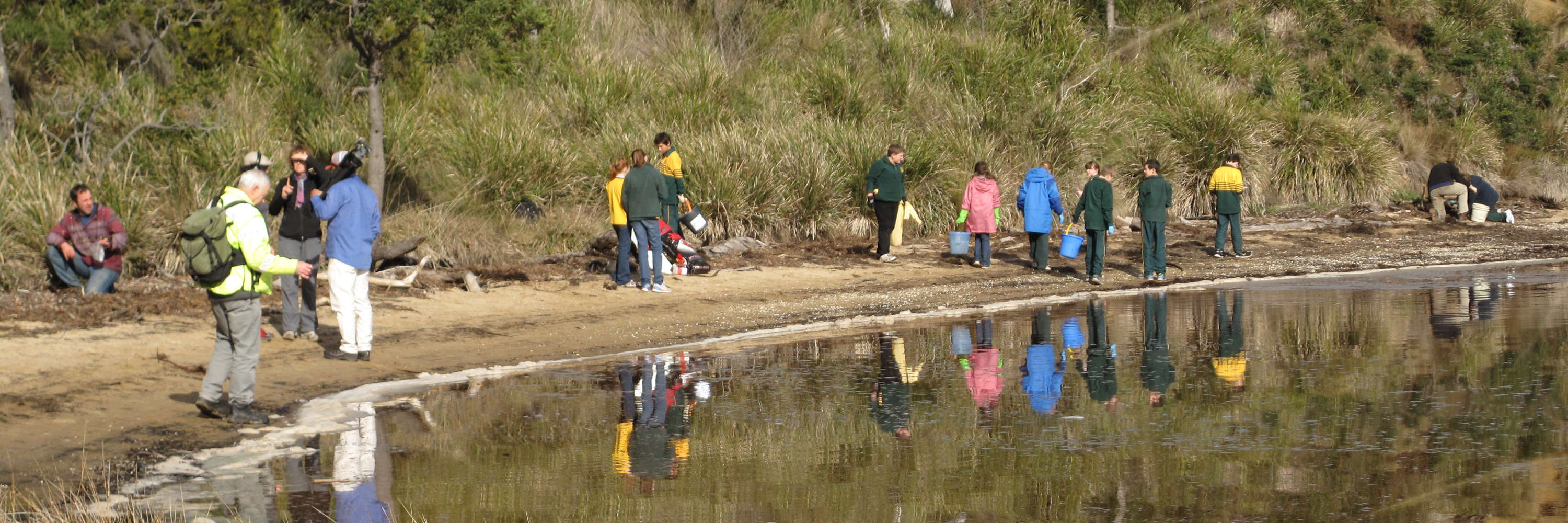 A group of people of all ages are scattered along a sandy water’s edge, surrounded by tall grass and reeds, collecting plastic in buckets. In the foreground, a child holds up a bottle. Photo: Andrew Hughes.