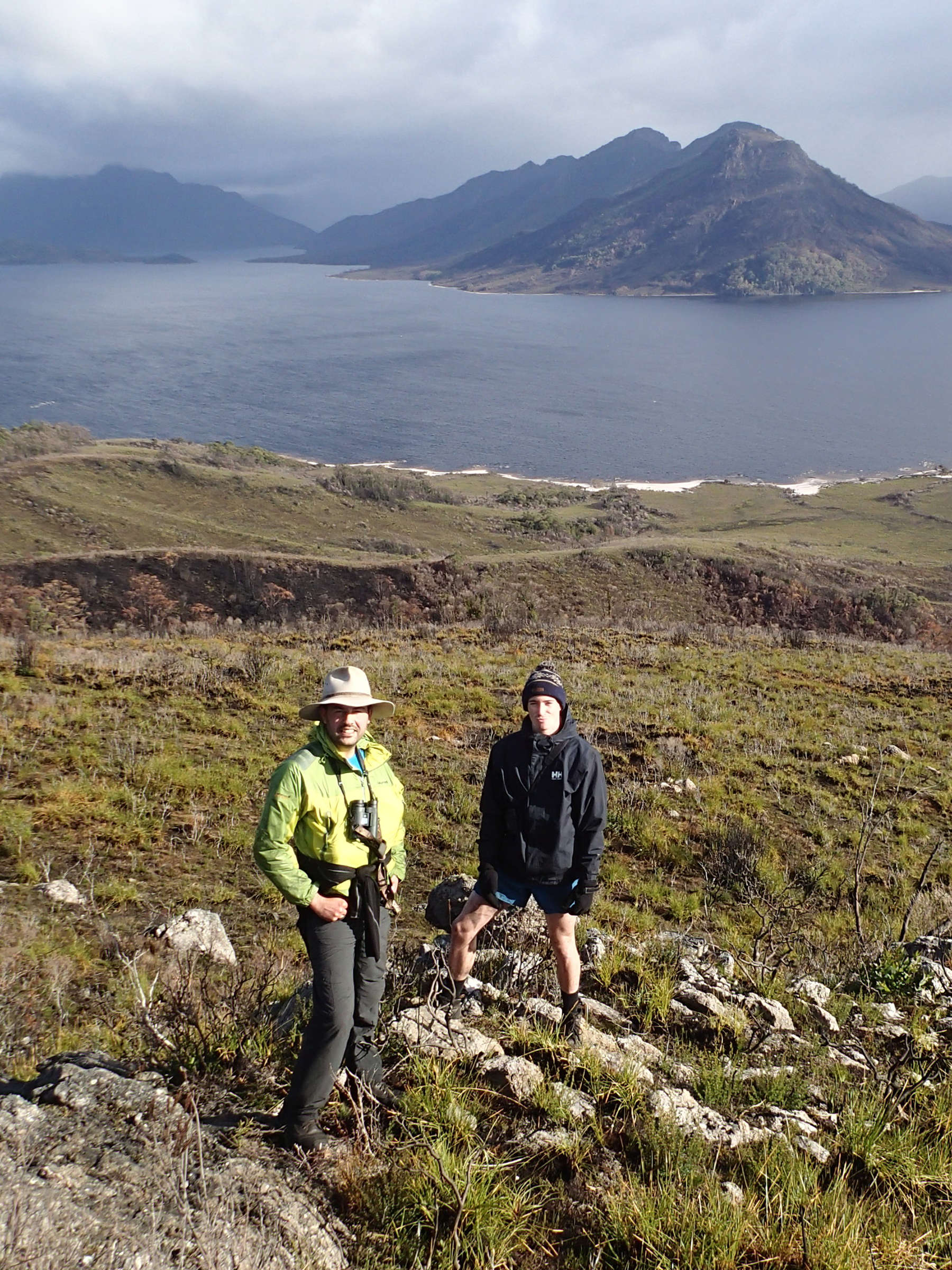 Two men standing on rocks surrounded by grassland, by a large lake in remote southwest Tasmania, with mountains on the far side of the lake. They are in the area doing Where? Where? Wedgie! surveys. They are smiling at the camera and one of them has a pair of binoculars. Photo: Nick Mooney.