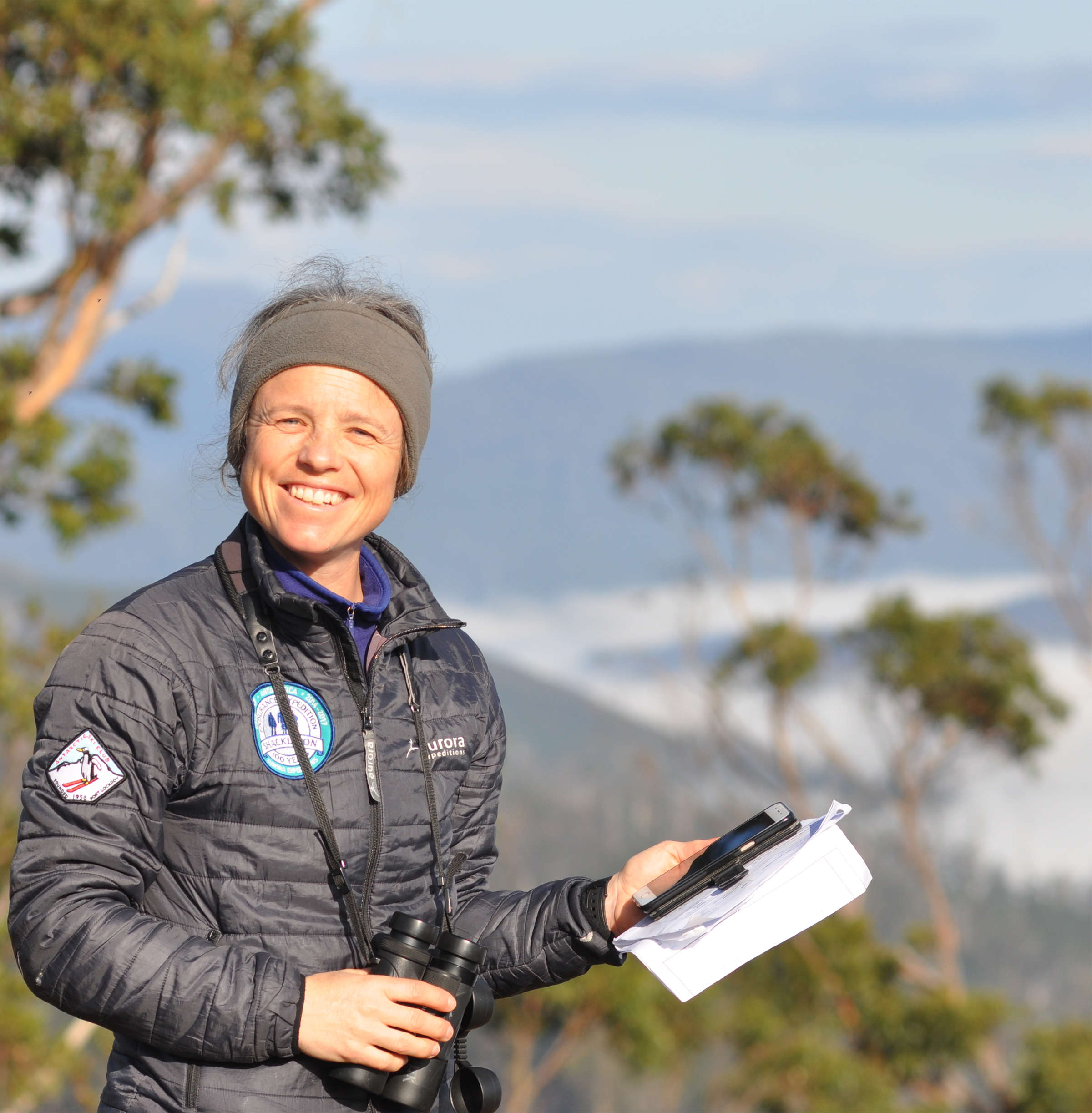 A woman wearing a down jacket and fleece headband smiles at the camera. She holds a smartphone and datasheets with one hand, while her other hand rests on her binoculars. Behind her are trees and, stretching off into the distance, a cloud-filled valley and mountains. Photo: Stephen Anstee.